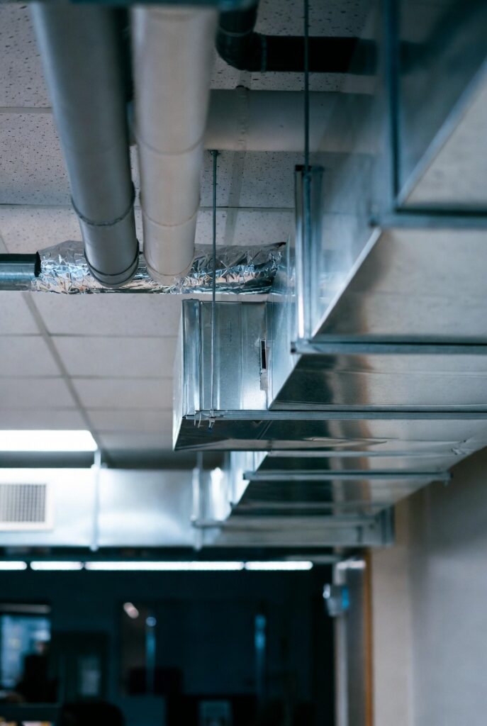 Silver metal HVAC ducts and ventilation pipes running along a commercial building ceiling with foil insulation and fluorescent lighting in the background