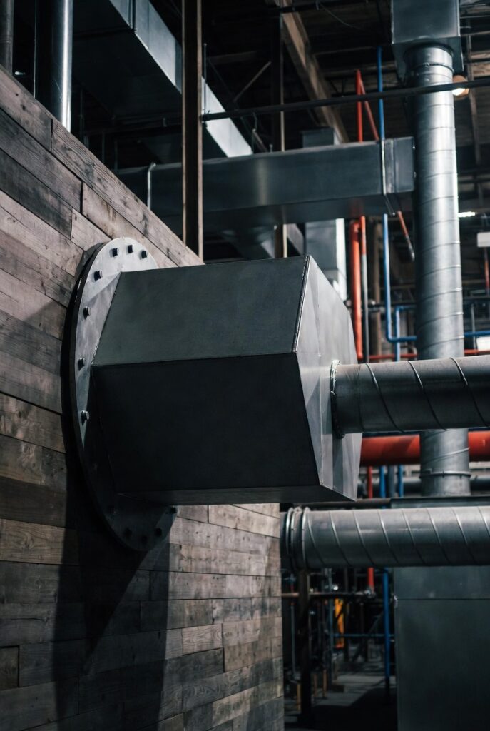 Close-up of a large industrial HVAC duct mounted through a wooden wall with metal pipes and ductwork visible in the background of a commercial building