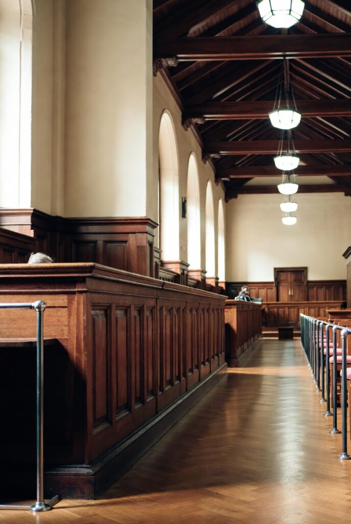 Wide interior view of a grand traditional courtroom with dark wooden paneling, high vaulted ceiling with exposed beams, arched windows, hanging pendant lights, wooden pews with pink cushioned public gallery seats, and a parquet hardwood floor