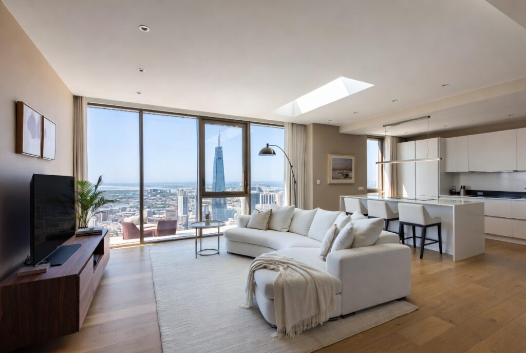 Spacious open-plan penthouse living room with white curved sofa, oak hardwood floors, floor-to-ceiling windows, and a sweeping city skyline view including a prominent skyscraper