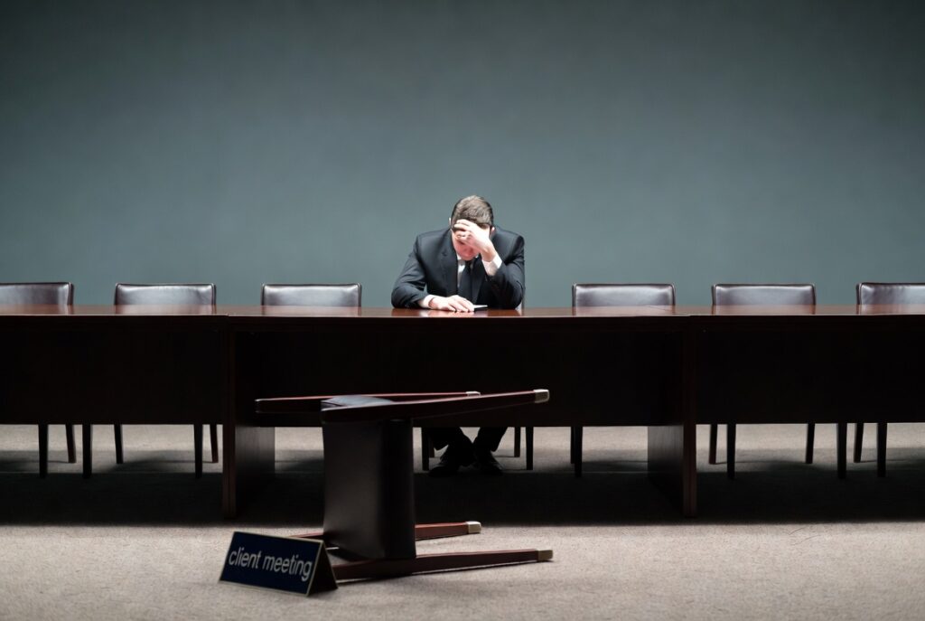 Businessman sitting alone at a long empty conference table with a fallen client meeting sign on the floor