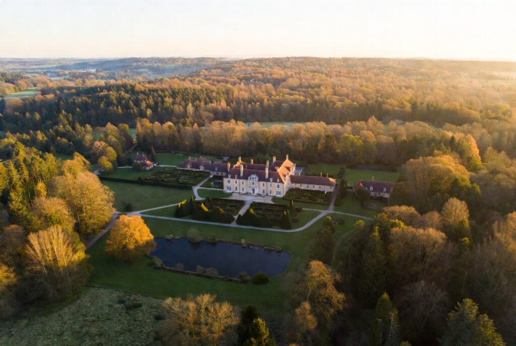 Aerial drone view of a grand English countryside estate with formal hedgerow gardens, a pond, and surrounding woodland at golden hour